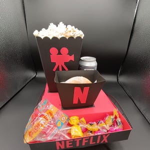 Puede incluir: Una caja roja y negra con la palabra "NETFLIX" impresa en la parte delantera. La caja contiene un recipiente de palomitas de maíz negro con un icono de cámara de cine rojo, un recipiente negro con una rosquilla blanca y la letra "N", una lata de refresco y una variedad de caramelos de colores.