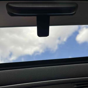 May include: A black car interior with a view of the sky through the sunroof. The sunroof has a black frame and a retractable sunshade. The sky is blue with white clouds.