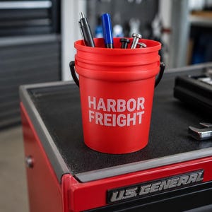 May include: A bright red plastic bucket with a black handle featuring the white text HARBOR FREIGHT printed on the front. The bucket contains a black pen, a blue marker, and several metal bolts and screws, serving as a tool storage container.