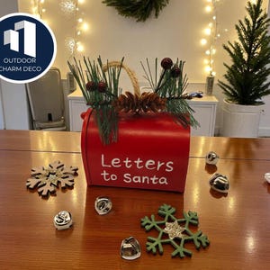 May include: A red mailbox with the text "Letters to Santa" is decorated with pine needles, pinecones, and berries. Silver bells and wooden snowflakes are scattered on the wooden table, creating a festive holiday scene.