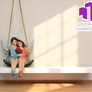 May include: A decorative figurine of a couple on a swing, with angel wings and holding a bouquet of red flowers. The couple is dressed in pastel colors. The swing is suspended by gold chains. The text "OUTDOOR CHARM DECO" is in the upper right corner.
