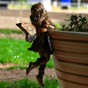 Puede incluir: Una figurita de hada de bronce con alas, sosteniendo una flor, posada en el borde de una maceta rayada.