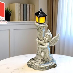 Op de afbeelding: Decoratieve buitenlamp met een cherubijn die een zuil vasthoudt met een zwart-gele lantaarn. De lamp is gemaakt van een grijs, steenachtig materiaal en staat op een ronde tafel. De tekst "Outdoor Charm Deco" staat in de linkerbovenhoek.