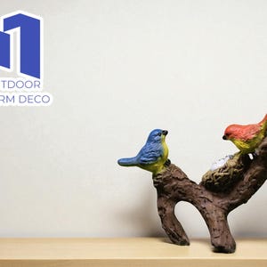 Może przedstawiać: Dekoracyjna rzeźba ptaków z dwoma kolorowymi ptakami siedzącymi na brązowej gałęzi z gniazdem. Ptaki są niebiesko-żółte i czerwono-żółte. Widoczny jest napis "OUTDOOR CHARM DECO".