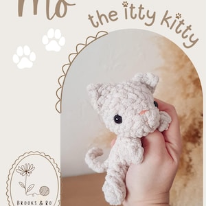 May include: A small, light beige crocheted cat plush toy with black plastic eyes and a stitched pink nose. The text 'Mo the itty kitty' appears at the top, with two white paw prints and a logo for 'Brooks & Ro' featuring a flower and yarn ball.