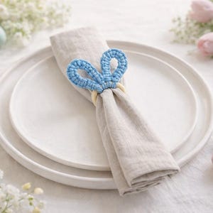 May include: A light beige linen napkin is held by a blue crochet bunny ear napkin ring, set on a white plate. Pastel Easter eggs and flowers surround the plate, creating a spring-themed tablescape.