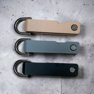 Op de afbeelding: Drie sleutelhangers met verschillende gekleurde metalen en kunststof behuizingen. De bovenste sleutelhanger is beige, de middelste sleutelhanger is lichtblauw en de onderste sleutelhanger is donkergrijs. Elke sleutelhanger heeft een zilveren ring en een zilveren knop.