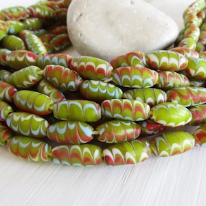 Puede incluir: Una colección de cuentas con patrones verdes, rojos, blancos y azules. Las cuentas de forma ovalada están ensartadas, creando un efecto en capas. Una piedra lisa de color claro descansa en el fondo.