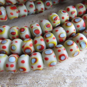 Op de afbeelding: Close-up van witte glazen kralen met rode, gele en blauwe patronen. De ovale kralen hebben een glanzende afwerking. De kralen zijn geregen, mogelijk voor een ketting of armband.