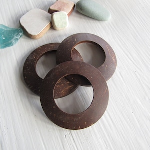 Puede incluir: Tres anillos de cáscara de coco marrón, apilados uno encima del otro. Los anillos están hechos de cáscara de coco natural y tienen un acabado liso y pulido.