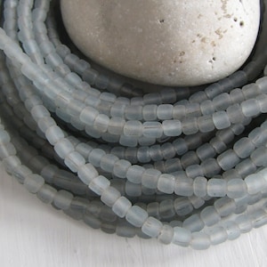 Può includere: Una collezione di perline di vetro azzurro con forma rettangolare arrotondata. Le perline sono infilate, formando più fili. Una pietra grigia liscia poggia sulle perline. Le perline sono probabilmente utilizzate per la creazione di gioielli.