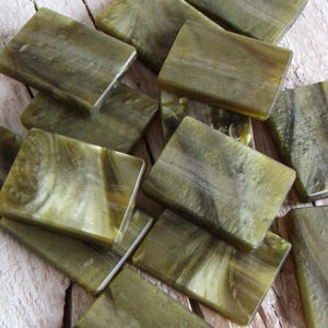 Può includere: Una collezione di piastrelle rettangolari verde oliva con aspetto marmorizzato. Le piastrelle sono disposte su una superficie di legno invecchiato, mostrando un'estetica naturale e organica. Finitura liscia e lucida.