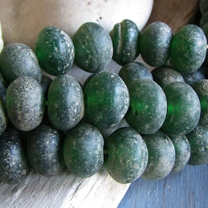 Puede incluir: Una hebra de cuentas de vidrio verde oscuro con una superficie texturizada y moteada. Las cuentas son redondas y tienen una forma ligeramente irregular.