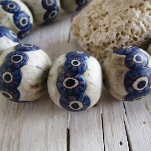 Peut inclure: Un collier de perles en céramique blanche avec des motifs peints bleus et rouges. Chaque perle présente un motif répété d'yeux stylisés.