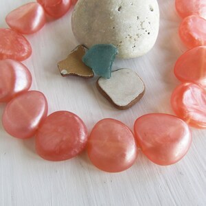 Può includere: Una collana realizzata con perline lisce a forma di ciottolo di colore corallo. Le perline sono disposte in cerchio e infilate su un cordoncino trasparente.