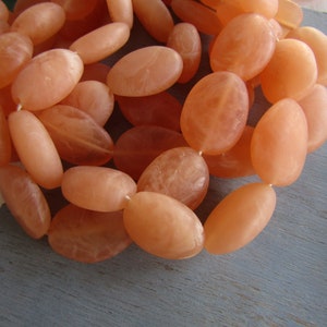 Puede incluir: Primer plano de un collar con cuentas ovaladas de color melocotón. Las cuentas están ensartadas, creando una forma fluida y orgánica. Las cuentas tienen una cualidad translúcida. El collar descansa sobre una superficie azul-gris.