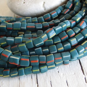 Può includere: Primo piano di una collana di perline quadrate turchesi con strisce colorate. Le perline hanno strisce bianche, gialle e arancioni. Le perline sono disposte su una superficie di legno bianco.