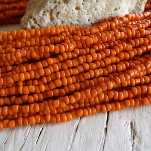 May include: A close-up of a strand of orange seed beads. The beads are small and round, and they are arranged in a tight, even pattern. The beads are strung on a clear thread, and they are illuminated by a soft, warm light.