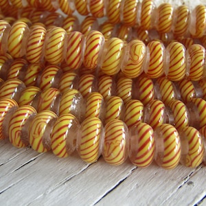 Op de afbeelding: Een streng gele en rode gestreepte glazen kralen. De kralen zijn rond en hebben een helder centrum. De kralen zijn geregen op een heldere draad.
