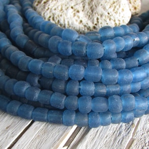 Può includere: Un filo di perline di vetro blu, ogni perlina è opaca e ha una forma leggermente arrotondata. Le perline sono disposte in un mucchio, mostrando la texture e il colore del vetro.