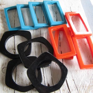 Può includere: Una collezione di componenti di gioielli geometrici e colorati. L'immagine presenta diversi pezzi rettangolari a telaio aperto in azzurro e arancione, insieme a pezzi neri di forma quadrata. I componenti sono disposti su una superficie di legno bianco invecchiato.