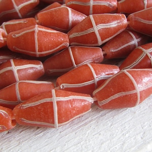 Op de afbeelding: Een streng oranje glazen kralen met witte strepen. De kralen zijn ovaalvormig en hebben een getextureerd oppervlak.