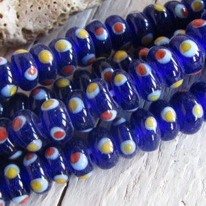 Op de afbeelding: Een streng kobaltblauwe glazen kralen met witte, gele en rode stippen. De kralen zijn rond en hebben een licht onregelmatige vorm.