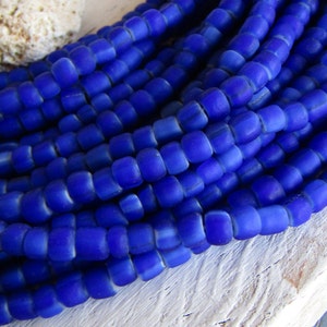 Op de afbeelding: Een close-up van een streng blauwe glazen kralen. De kralen zijn klein en rond, met een licht gematteerde afwerking. Ze zijn in een losse stapel gerangschikt, waardoor de individuele kralen zichtbaar zijn.