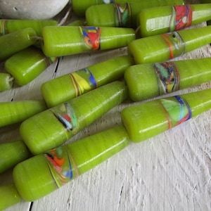 Op de afbeelding: Een verzameling groene glazen kralen met kleurrijke swirls en strepen. De kralen zijn lang en smal, met een puntig uiteinde. Ze zijn gerangschikt op een wit houten oppervlak.