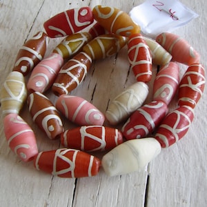 Può includere: Una collezione di perline ovali colorate con disegni bianchi a spirale. Le perline sono in tonalità di rosso, rosa, marrone e giallo e sono infilate insieme. Lo sfondo è una superficie di legno bianco e invecchiato.