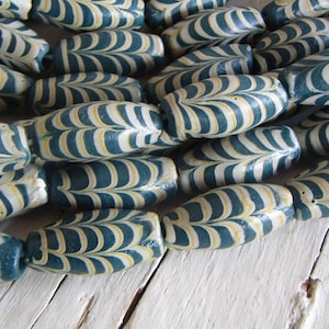 Pode incluir: Close-up de uma coleção de contas listradas azuis, brancas e amarelas. As contas são ovais e têm um padrão semelhante a uma folha. As contas estão dispostas próximas umas das outras, criando uma superfície texturizada.