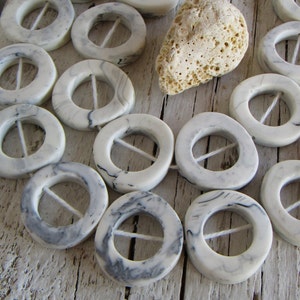 Può includere: Una collana di bottoni rotondi marmorei bianchi e grigi con un foro al centro e una linea bianca che attraversa il foro. I bottoni sono disposti in cerchio su una superficie di legno bianco.