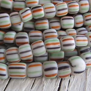 Puede incluir: Primer plano de una hebra de cuentas de vidrio blanco con rayas naranjas, verdes y negras. Las cuentas están dispuestas en un patrón aleatorio.