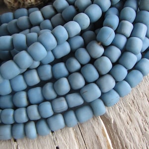Può includere: Un filo di perline di vetro blu chiaro. Le perline sono piccole e rotonde, con una finitura opaca. Sono disposte in un mucchio, mostrando la trama delle perline.