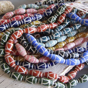 Op de afbeelding: Een verzameling kleurrijke glazen kralen met witte geometrische patronen. De kralen zijn ovaalvormig en hebben een glanzende afwerking.