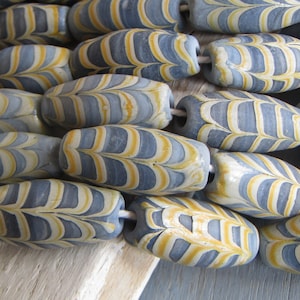 Puede incluir: Primer plano de un collar de cuentas hechas a mano. Cada cuenta es de color azul grisáceo claro con patrones amarillos y blancos en forma de hoja. Las cuentas están ensartadas con un hilo blanco, creando un diseño único y artístico.