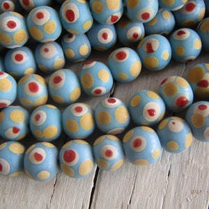 May include: A collection of light blue wooden beads with a pattern of yellow and red dots. The beads are round and appear to be handmade, with a slightly rustic aesthetic. The beads are scattered across a white wooden surface.