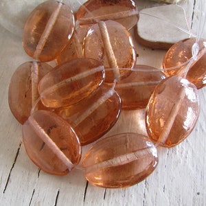 Può includere: Una collezione di perline ovali traslucide in un caldo colore pesca. Ogni perlina presenta un design lineare bianco che la attraversa al centro. Le perline sono infilate su un filo trasparente, suggerendo che siano per la creazione di gioielli.