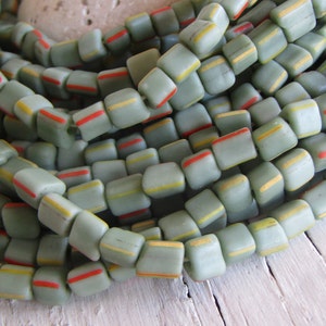 Può includere: Una collana di perline quadrate in vetro con una base di colore verde chiaro. Le perline hanno sottili strisce di vernice gialla e rossa.