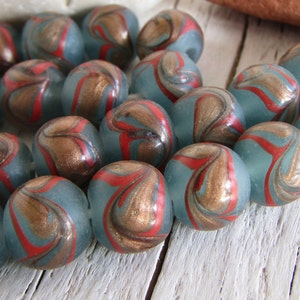 Puede incluir: Una hebra de cuentas de vidrio azul verdoso con patrones de remolino rojo y dorado. Las cuentas son redondas y tienen un acabado brillante.