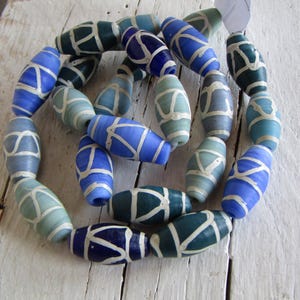 Può includere: Una collana di perline ovali nei toni del blu e del verde, con un motivo geometrico bianco. Le perline sono infilate insieme, creando una forma circolare. Le perline sono su una superficie di legno bianco.