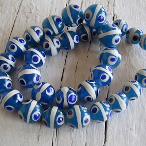 Op de afbeelding: Een streng blauwe glazen kralen met witte strepen en oogvormige patronen. De kralen zijn rond en aan elkaar geregen, waardoor een cirkelvorm ontstaat. De kralen liggen op een wit oppervlak.
