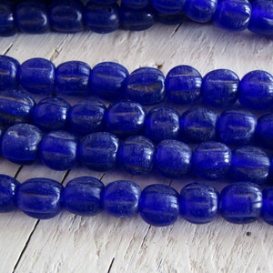 Op de afbeelding: Een streng diepblauwe glazen kralen met een licht ovale vorm. De kralen hebben een getextureerd oppervlak en zijn aan elkaar geregen op een doorzichtige draad.