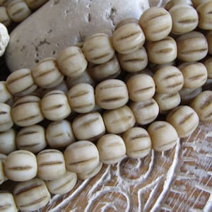 Op de afbeelding: Een streng ovale, gebroken witte kralen met een gegroefd patroon. De kralen zijn aan elkaar geregen op een wit koord.