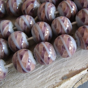 Op de afbeelding: Een streng ambachtelijke glazen kralen. Elke kraal heeft een afgeronde vorm met een wervelend patroon van lavendel, perzik en wit. De kralen zijn geregen op een wit koord en zijn geschikt voor het maken van sieraden.