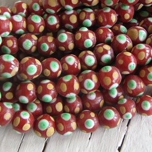 May include: Close-up of a collection of red beads with a glossy finish. Each bead features a pattern of yellow and green spots. The beads are round and appear to be strung together, suitable for jewelry making or crafting.