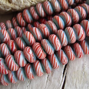 Könnte beinhalten: Eine Strähne aus blauen und weißen Glasperlen mit roten Streifen. Die Perlen sind rund und haben eine leicht unregelmäßige Form. Sie sind auf einem durchsichtigen Faden aufgefädelt.