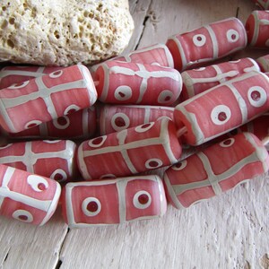 Op de afbeelding: Een streng roze en witte glazen kralen met een geometrisch patroon. De kralen zijn cilindrisch en hebben een wit rasterpatroon met witte cirkels op een roze achtergrond.