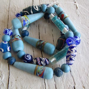 Op de afbeelding: Een verzameling handgemaakte glazen kralen in verschillende blauwtinten. De kralen omvatten cilindrische, kegelvormige en ronde ontwerpen, met decoratieve patronen in wit, rood en geel. De kralen zijn aan elkaar geregen, waardoor een ketting of armband ontstaat.