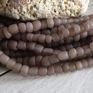 Könnte beinhalten: Nahaufnahme von Strängen aus hellvioletten, zylindrischen Perlen. Die Perlen sind in mehreren Reihen angeordnet, mit einem rauen, beigen Stein im Hintergrund. Die Perlen haben ein leicht durchscheinendes Aussehen, was darauf hindeutet, dass sie aus Glas oder einem ähnlichen Material bestehen. Das Thema ist Schmuck.